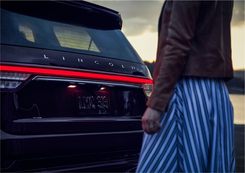 A person is shown near the rear of a 2023 Lincoln Aviator® SUV as the Lincoln Embrace illuminates the rear lights | Central Florida Lincoln in Orlando FL